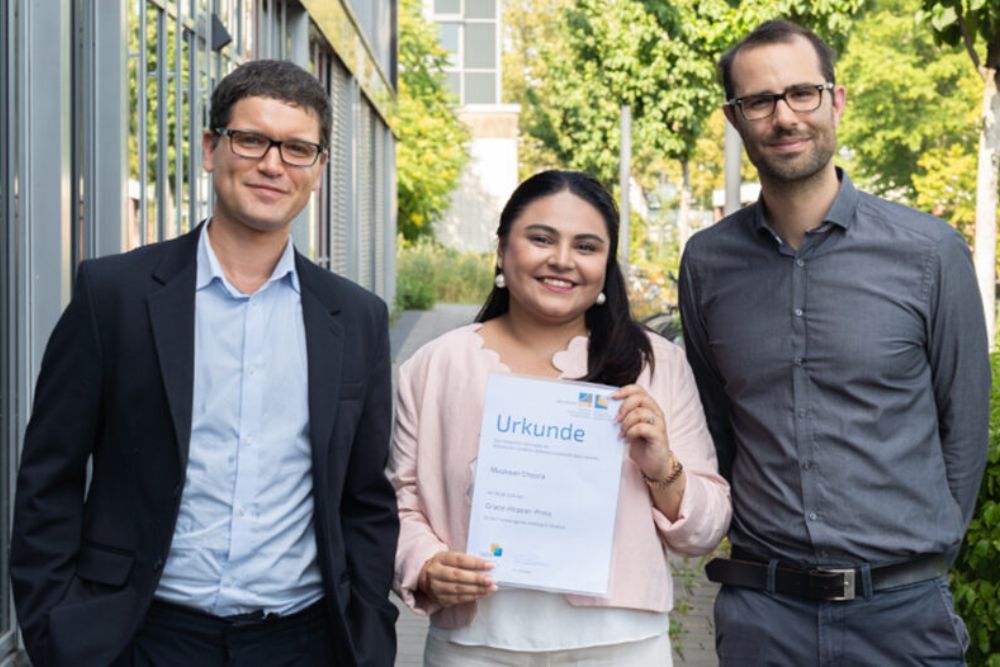 Muskaan Chopra holds her Grace Hopper Award certificate outside a University of Bonn building, accompanied by her supervisors Prof. Dr. Rafet Sifa and Dr. Lorenz Sparrenberg.