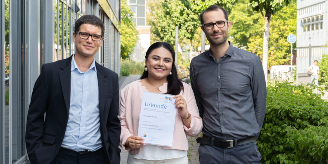 Muskaan Chopra holds her Grace Hopper Award certificate outside a University of Bonn building, accompanied by her supervisors Prof. Dr. Rafet Sifa and Dr. Lorenz Sparrenberg.