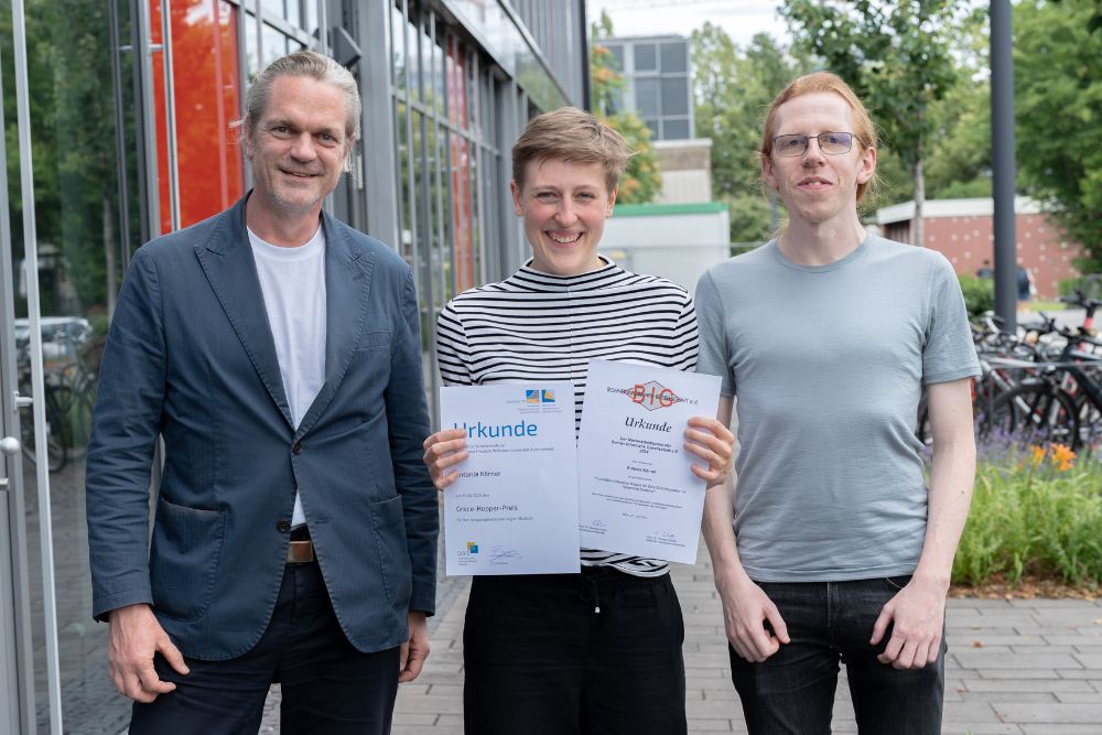 Three people are standing in front of a university building. On the left is Prof. Dr. Christian Bauckhage, in the middle is Grace Hopper Award winner Antonia Körner, proudly holding her certificates in the picture. On the right is Patrick Seifner, PhD candidate at the Lamarr Institute and Antonia Körner's supervisor.