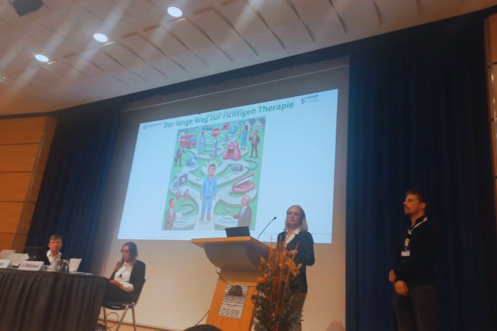 Dr. rer. medic. Sarah Dietz-Terjung and Sebastian Buschjäger present the Sleepwalker tool at the DGSM annual conference. In the background is a slide entitled 'The long road to the right therapy', which graphically depicts the therapeutic process. Both are standing at a lectern in a conference hall with blue curtains.