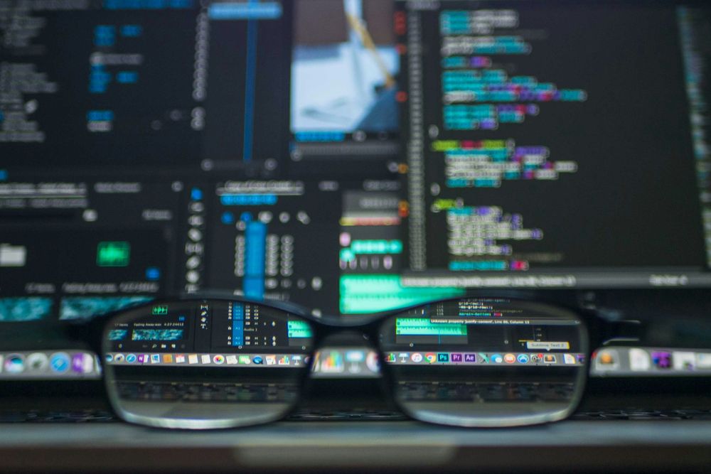 A pair of eyeglasses resting on a laptop keyboard, with the laptop screen displaying lines of colorful code and data visualizations. The view through the glasses is sharp and focused on the screen content, while the surrounding area appears blurred.
